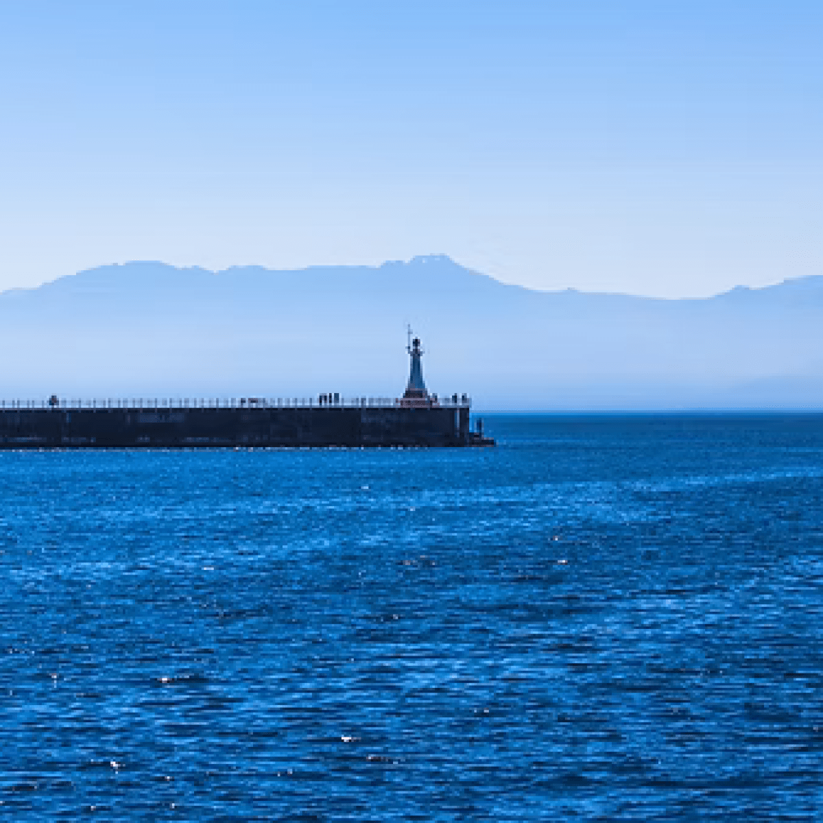 Blue ocean view with distant pier and mountains under clear sky.
